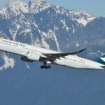 A Cathay Pacific aircraft is captured mid-flight against a backdrop of snow-capped mountains. The aeroplane, with its landing gear partially retracted, is ascending into the clear blue sky.