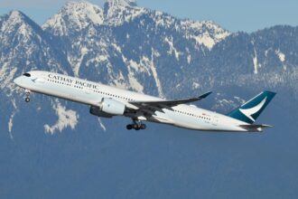 A Cathay Pacific aircraft is captured mid-flight against a backdrop of snow-capped mountains. The aeroplane, with its landing gear partially retracted, is ascending into the clear blue sky.