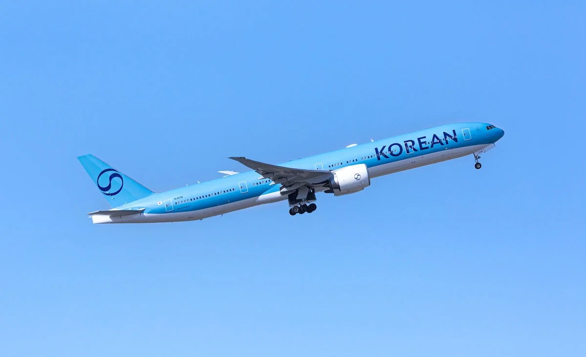 A Korean Air airplane, painted in light blue with the word "KOREAN" prominently displayed on its fuselage, is captured mid-flight against a clear blue sky. The aircraft's landing gear is still extended as it ascends.