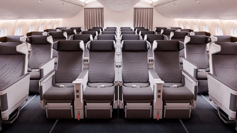 The interior of an airplane cabin showing rows of premium economy seats. The seats are arranged in a 2-4-2 configuration, with ample legroom and footrests. The cabin is well-lit with overhead lighting, and the seats have a modern design with dark gray upholstery and white armrests.