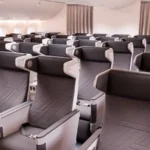 The image shows the interior of an airplane cabin with rows of premium economy seats. The seats are arranged in a 2-4-2 configuration, featuring grey upholstery with black headrests and armrests. Each seat has a personal entertainment screen, USB ports, and power outlets. The cabin is well-lit with natural light coming through the windows on the left side.