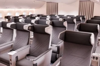 The image shows the interior of an airplane cabin with rows of premium economy seats. The seats are arranged in a 2-4-2 configuration, featuring grey upholstery with black headrests and armrests. Each seat has a personal entertainment screen, USB ports, and power outlets. The cabin is well-lit with natural light coming through the windows on the left side.