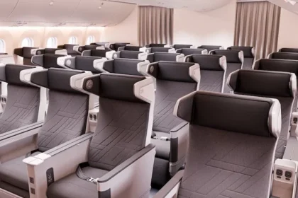 The image shows the interior of an airplane cabin with rows of premium economy seats. The seats are arranged in a 2-4-2 configuration, featuring grey upholstery with black headrests and armrests. Each seat has a personal entertainment screen, USB ports, and power outlets. The cabin is well-lit with natural light coming through the windows on the left side.