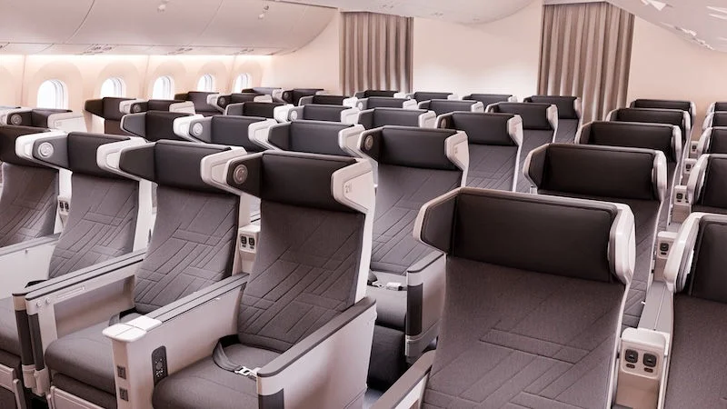 The image shows the interior of an airplane cabin with rows of premium economy seats. The seats are arranged in a 2-4-2 configuration, featuring grey upholstery with black headrests and armrests. Each seat has a personal entertainment screen, USB ports, and power outlets. The cabin is well-lit with natural light coming through the windows on the left side.