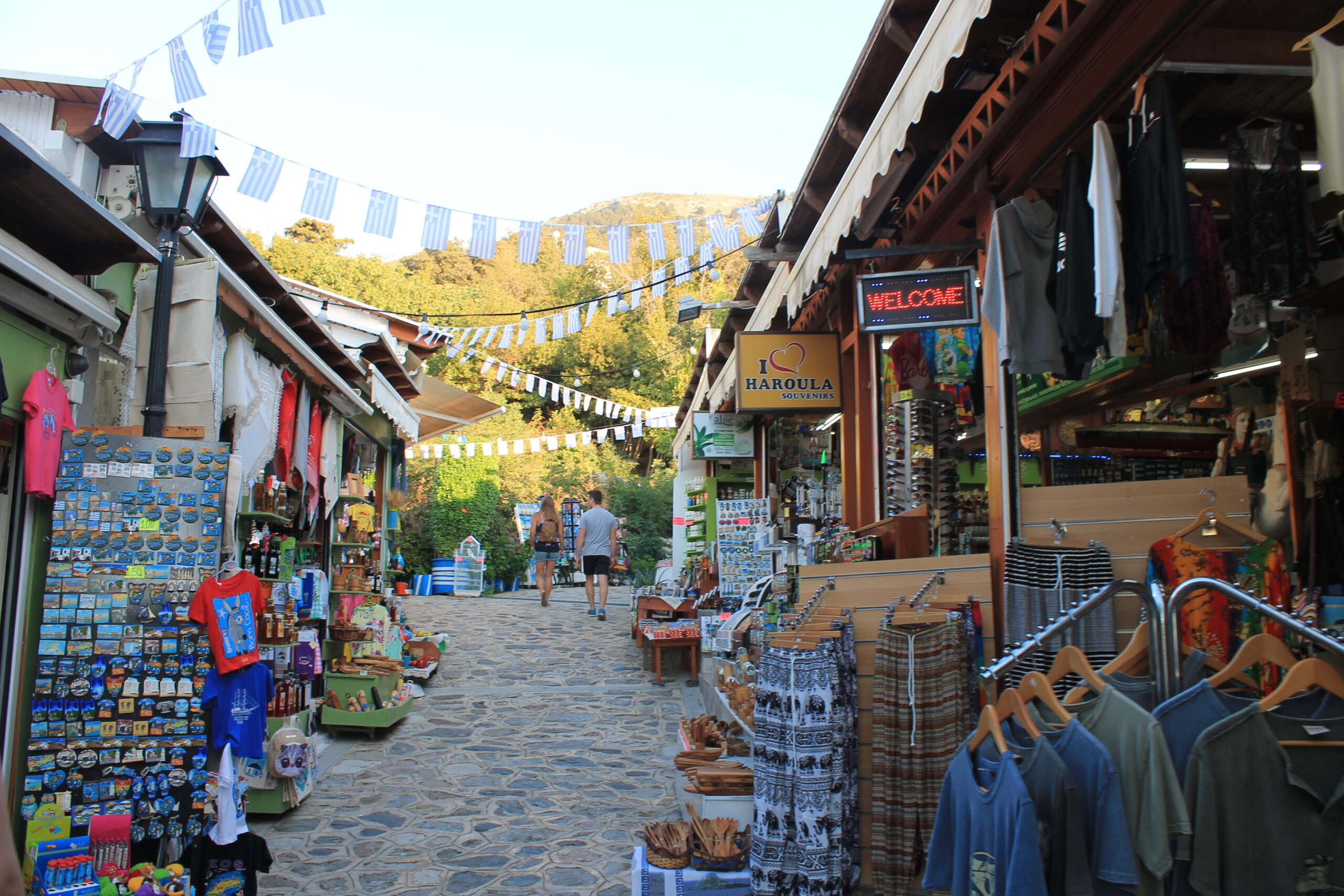 A paved street in Kos, with tourist shops selling a wide variety of souvenirs on either side, with goods placed along the street.