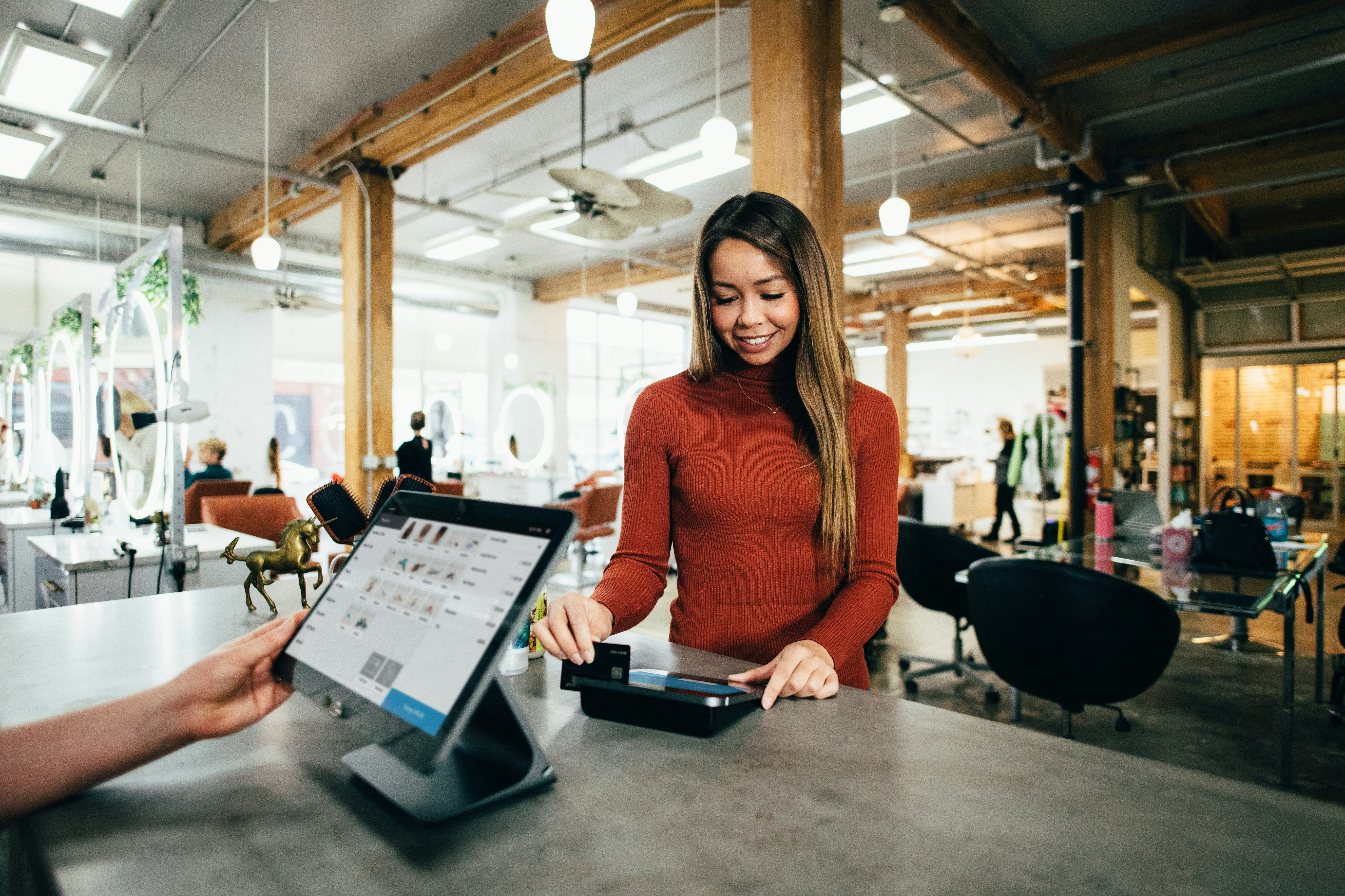  A woman in a red sweater is making a payment using a credit card at a modern, well-lit salon. The cashier's hand is holding a tablet displaying the payment interface. The salon has wooden beams, large windows, and several styling stations with circular mirrors and chairs.