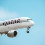 A Qatar Airways airplane is captured mid-flight against a clear blue sky. The aircraft's landing gear is extended, indicating it is either taking off or preparing to land. The airline's name "QATAR" is prominently displayed on the side of the plane in bold maroon letters, along with Arabic script.