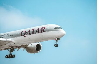 A Qatar Airways airplane is captured mid-flight against a clear blue sky. The aircraft's landing gear is extended, indicating it is either taking off or preparing to land. The airline's name "QATAR" is prominently displayed on the side of the plane in bold maroon letters, along with Arabic script.