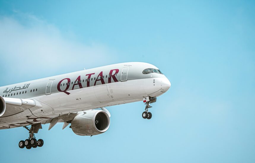 A Qatar Airways airplane is captured mid-flight against a clear blue sky. The aircraft's landing gear is extended, indicating it is either taking off or preparing to land. The airline's name "QATAR" is prominently displayed on the side of the plane in bold maroon letters, along with Arabic script.
