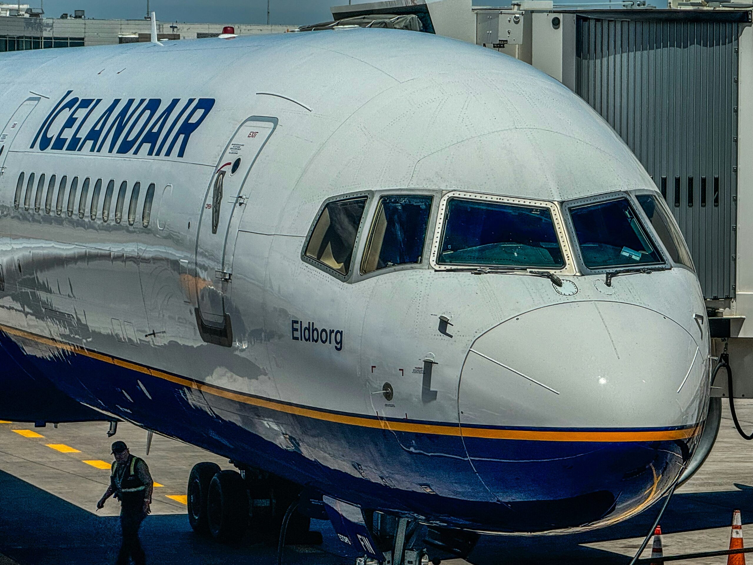 A close-up view of the front section of an Icelandair aeroplane parked at a gate. The aircraft is connected to the jet bridge on the right side. A ground crew member in a reflective vest is walking near the nose wheel of the plane.
