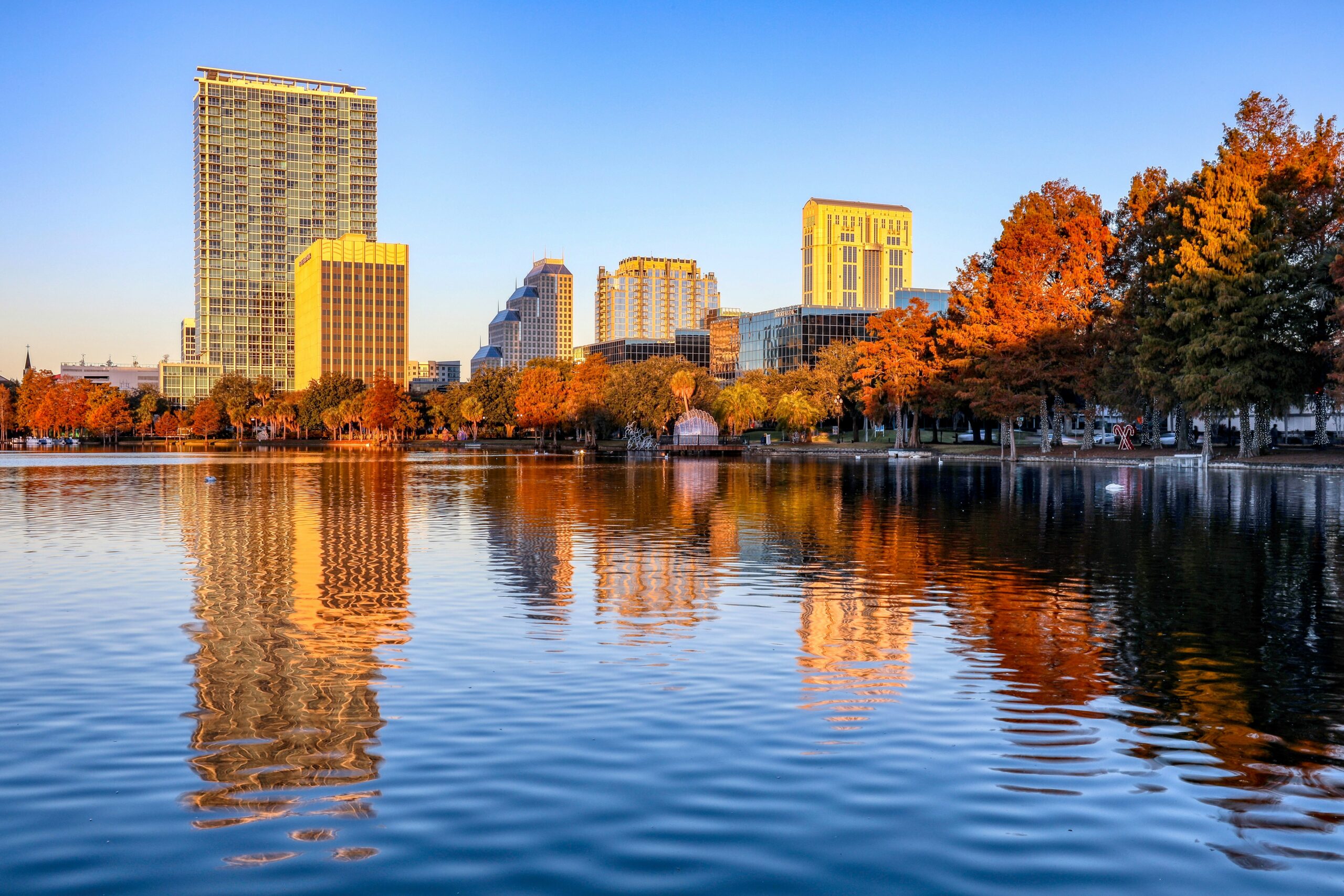 A scenic view of downtown Orlando, Florida, featuring tall buildings reflecting in the calm waters of Lake Eola during sunset. The trees along the lake's edge display vibrant autumn colours.