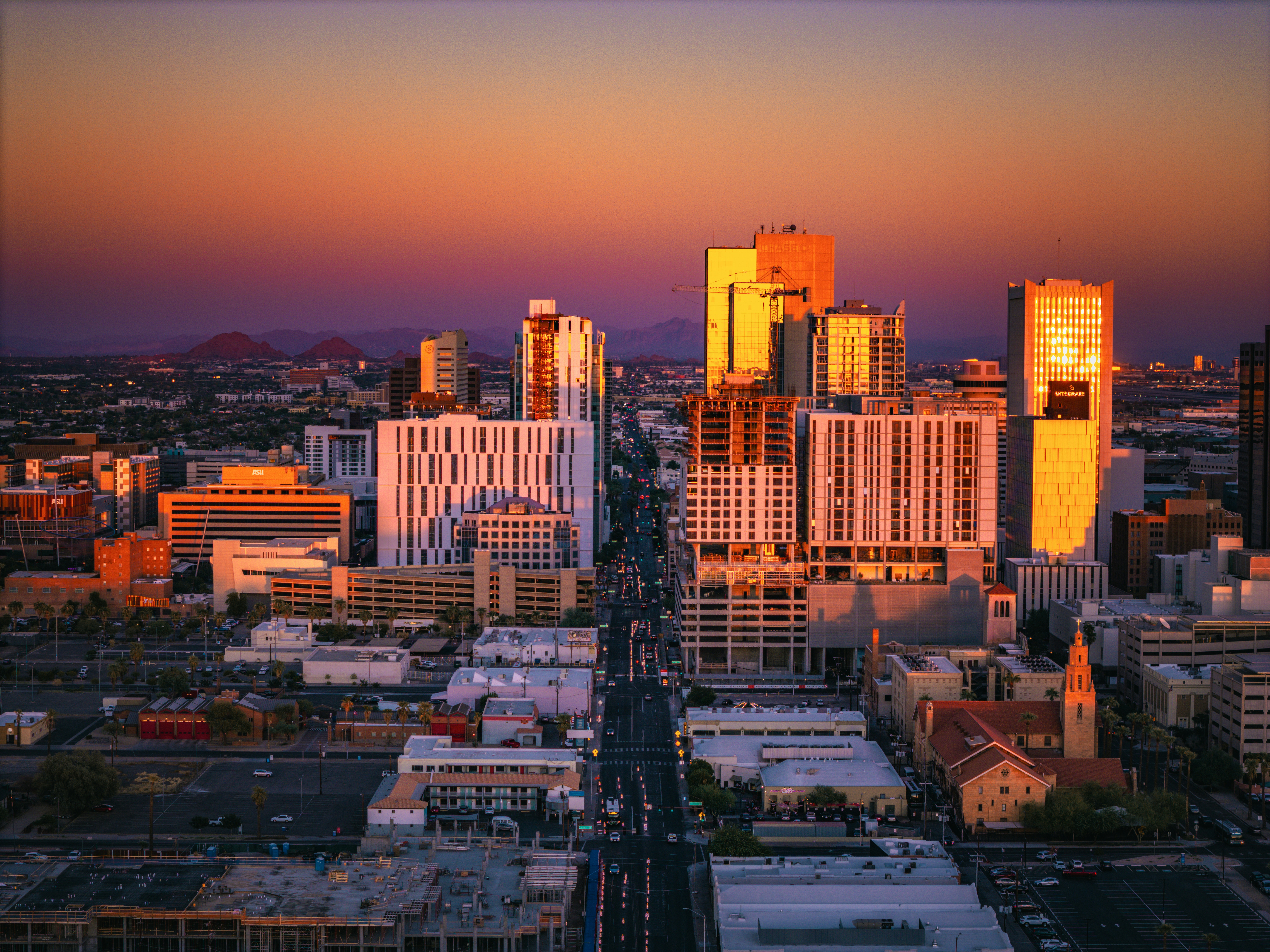 Aerial view of downtown Phoenix, Arizona at sunset. The cityscape features a mix of modern high-rise buildings and older structures, with the tallest buildings reflecting the golden hues of the setting sun. The streets are lined with cars and various smaller buildings, while mountains can be seen in the distant background under a gradient sky transitioning from orange to purple.