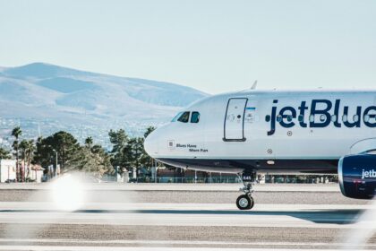 A jetBlue airplane is taxiing on the runway with a mountainous landscape in the background. The aircraft's nose and front landing gear are visible, along with the text "Blues Have More Fun" written near the cockpit. The plane's registration number "645" is also visible on the landing gear.