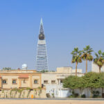 A view of the Al Faisaliyah Center, a prominent skyscraper in Riyadh, Saudi Arabia, with its distinctive pyramid shape and golden glass sphere near the top. In the foreground are residential buildings and palm trees under a clear blue sky.
