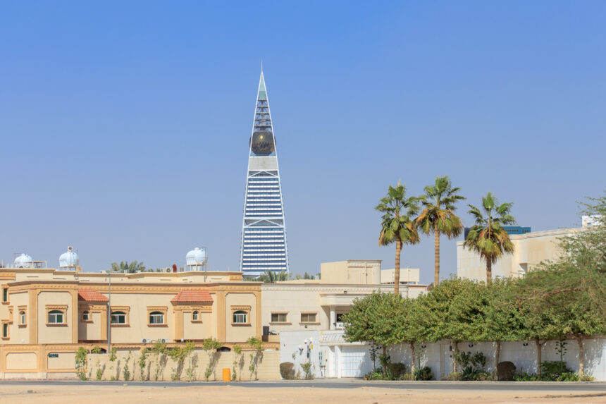 A view of the Al Faisaliyah Center, a prominent skyscraper in Riyadh, Saudi Arabia, with its distinctive pyramid shape and golden glass sphere near the top. In the foreground are residential buildings and palm trees under a clear blue sky.