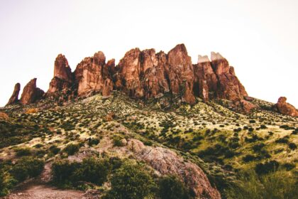 A rugged mountain range with steep, jagged peaks rises prominently against a clear sky. The foreground features a rocky terrain interspersed with patches of green vegetation and small shrubs.