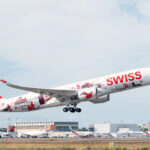 A Swiss International Air Lines aircraft, adorned with red and white livery featuring Swiss cultural symbols and landmarks, takes off from an airport. The plane's tail displays a large white cross on a red background, representing the Swiss flag. Several other airplanes and airport buildings are visible in the background under a partly cloudy sky.
