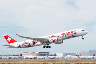 A Swiss International Air Lines aircraft, adorned with red and white livery featuring Swiss cultural symbols and landmarks, takes off from an airport. The plane's tail displays a large white cross on a red background, representing the Swiss flag. Several other airplanes and airport buildings are visible in the background under a partly cloudy sky.