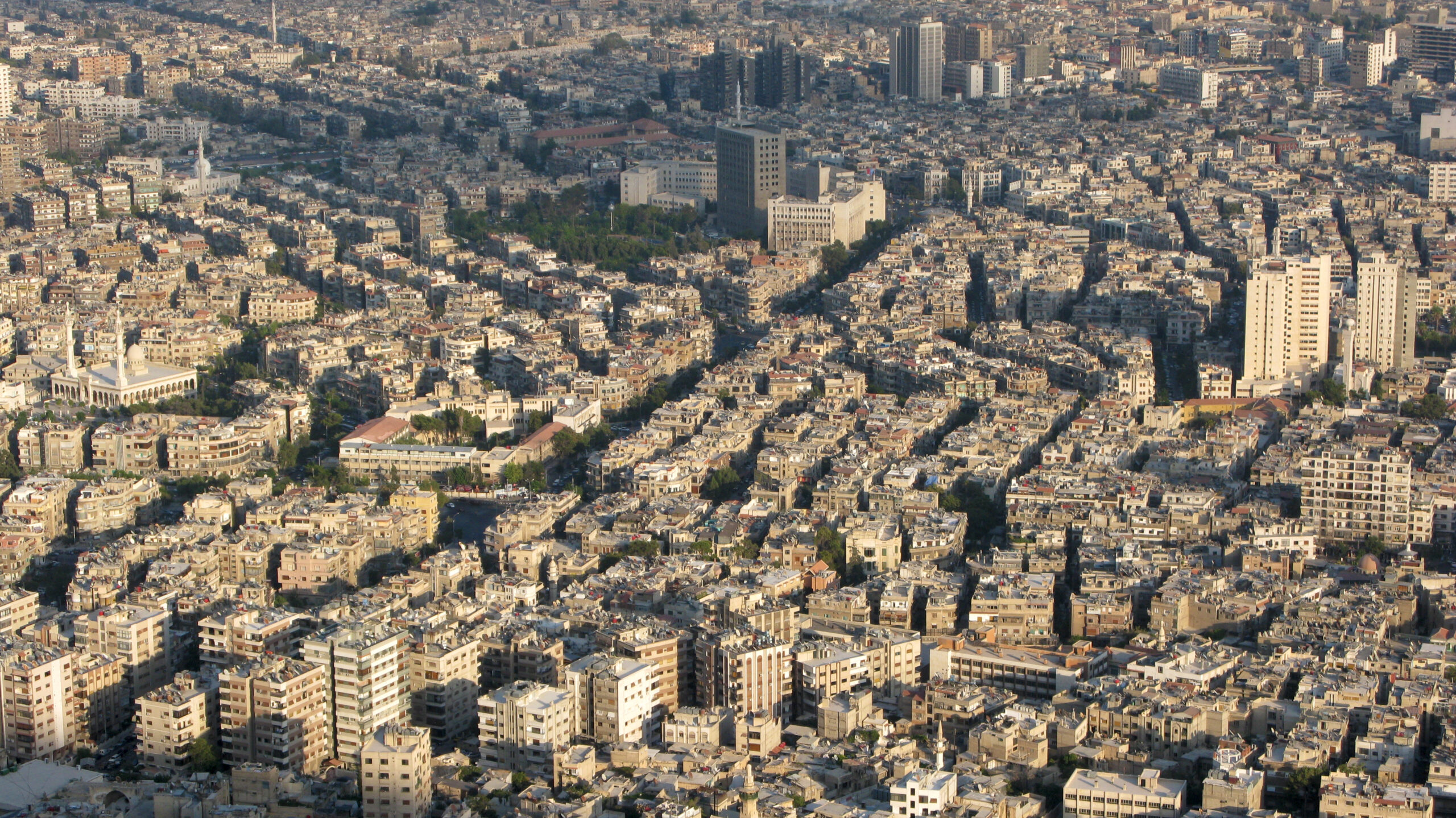 A panorama of the city of Damascus at sunset, with many sand-coloured apartment buildings reflecting the sunlight.