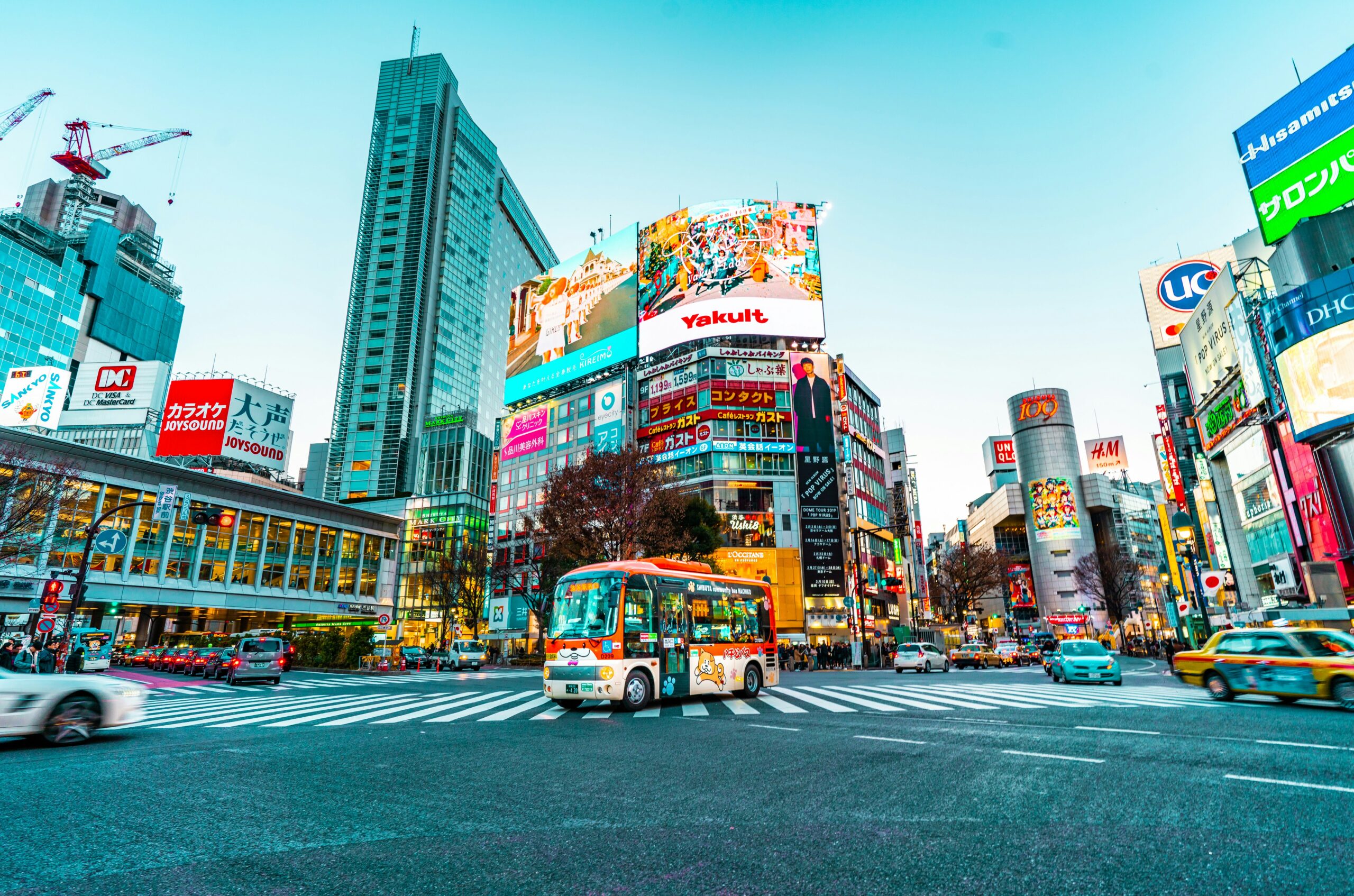 A bustling city intersection in Shibuya, Tokyo, featuring a colorful bus with cartoon characters on it crossing the street. Surrounding the intersection are tall buildings adorned with numerous bright advertisements and billboards, including prominent signs for Yakult, JOYSOUND, and H&M. The sky is clear and blue, adding to the vibrant atmosphere of the scene.
