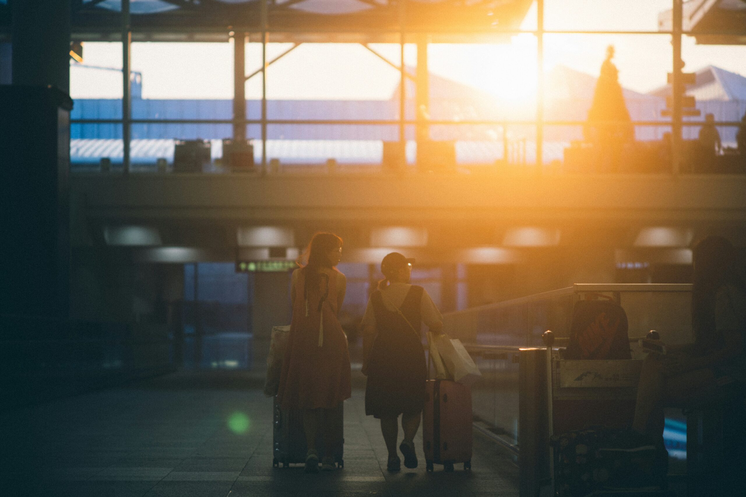 Two people walking through an airport terminal with luggage, illuminated by the warm glow of the setting sun streaming through large windows.