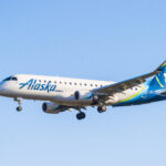 An Alaska Airlines aircraft is captured in mid-air against a clear blue sky. The aeroplane has its landing gear extended, indicating it is either approaching for landing or has just taken off. The aircraft features the Alaska Airlines logo prominently on its fuselage and a stylised image of a mountain on its tail.