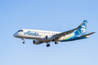 An Alaska Airlines aircraft is captured in mid-air against a clear blue sky. The aeroplane has its landing gear extended, indicating it is either approaching for landing or has just taken off. The aircraft features the Alaska Airlines logo prominently on its fuselage and a stylised image of a mountain on its tail.
