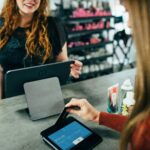 person swipes a credit card on a payment terminal at a retail counter, with various products and shelves in the background. The screen shows an amount of $102.49 and prompts to "Insert, Tap or Swipe."