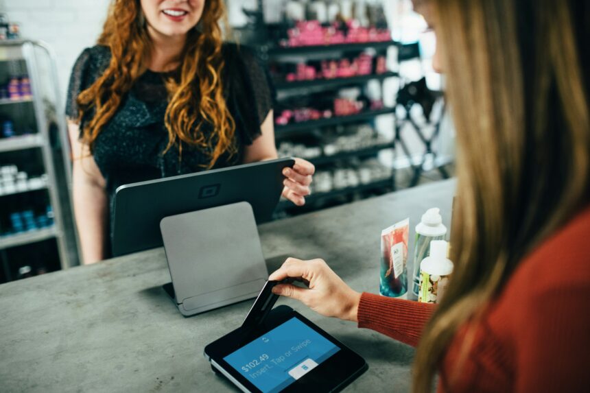 person swipes a credit card on a payment terminal at a retail counter, with various products and shelves in the background. The screen shows an amount of $102.49 and prompts to "Insert, Tap or Swipe."