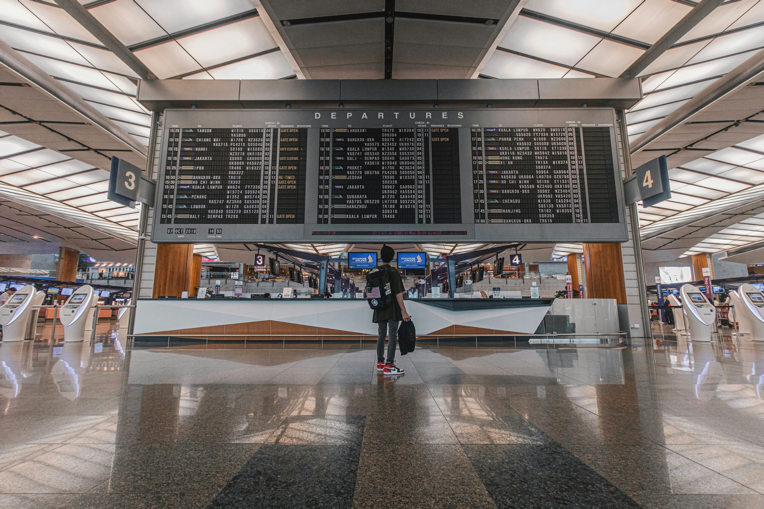 A traveler stands in front of a large departures board at an airport, looking up at the flight information. The board displays various flights, destinations, and statuses. The airport terminal is spacious with high ceilings and check-in kiosks on either side.