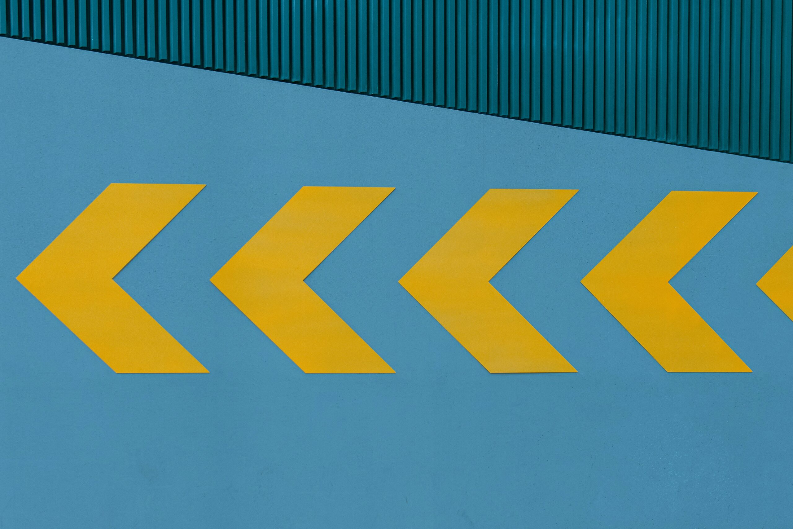 A blue wall with four large yellow left-pointing arrows in a row. Above the wall, there is a section of green corrugated metal.