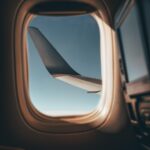 View from an airplane window showing the aircraft’s wing against a clear blue sky, with soft sunlight illuminating the window frame and part of the cabin interior.