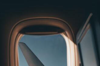 View from an airplane window showing the aircraft’s wing against a clear blue sky, with soft sunlight illuminating the window frame and part of the cabin interior.