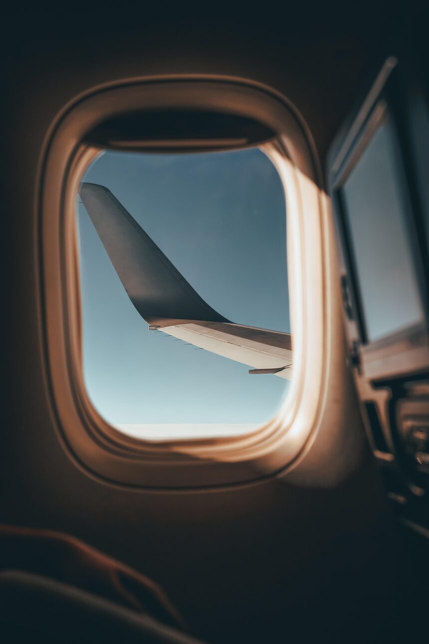 View from an airplane window showing the aircraft’s wing against a clear blue sky, with soft sunlight illuminating the window frame and part of the cabin interior.