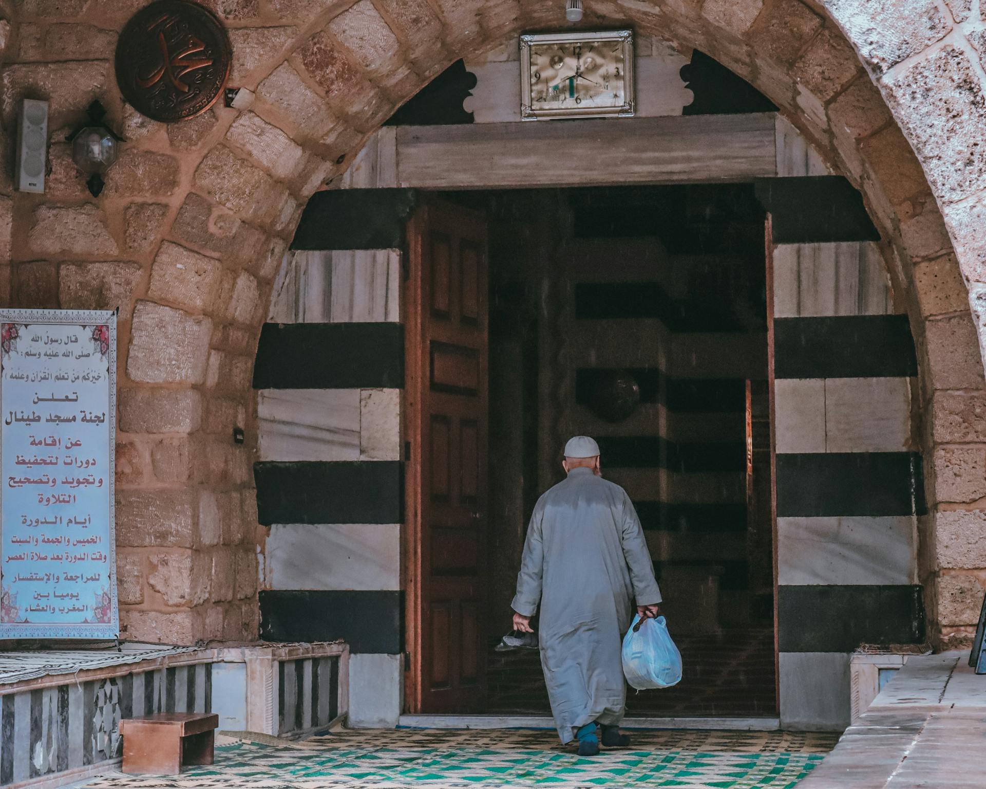 A man holding a carrier bag, walking towards the entrance of a mosque. His back is turned to the camera.