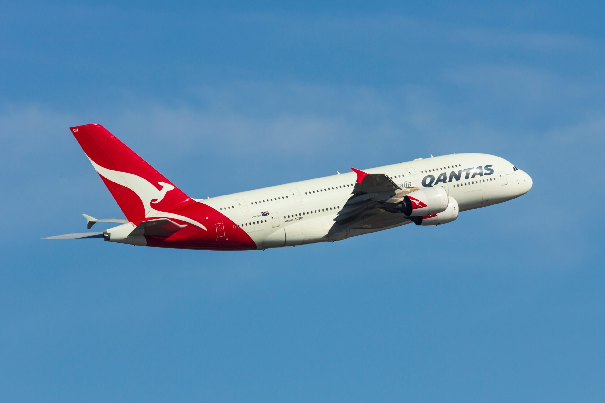 A Qantas aircraft mid-flight with the photo taken from a sideways angle. The plane appears in front of a blue sky background.