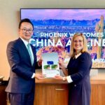 China Airlines President Kevin Chen and Phoenix Mayor Kate Gallego stand side by side holding an open folder with documents, in front of a screen that reads “PHOENIX WELCOMES CHINA AIRLINES” with a desert landscape background.