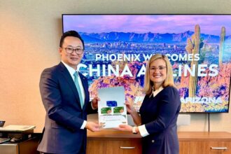 China Airlines President Kevin Chen and Phoenix Mayor Kate Gallego stand side by side holding an open folder with documents, in front of a screen that reads “PHOENIX WELCOMES CHINA AIRLINES” with a desert landscape background.