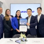 A group of five individuals in business attire stand together in a corporate setting, holding a certificate. The background features the Boeing logo prominently displayed on a screen. Two nameplates are visible on the table in front of them, reading "Crystal Remfert - Boeing Global Services" and "Chan Woo Jung - Korean Air." A floral arrangement is also present on the table.