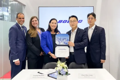 A group of five individuals in business attire stand together in a corporate setting, holding a certificate. The background features the Boeing logo prominently displayed on a screen. Two nameplates are visible on the table in front of them, reading "Crystal Remfert - Boeing Global Services" and "Chan Woo Jung - Korean Air." A floral arrangement is also present on the table.