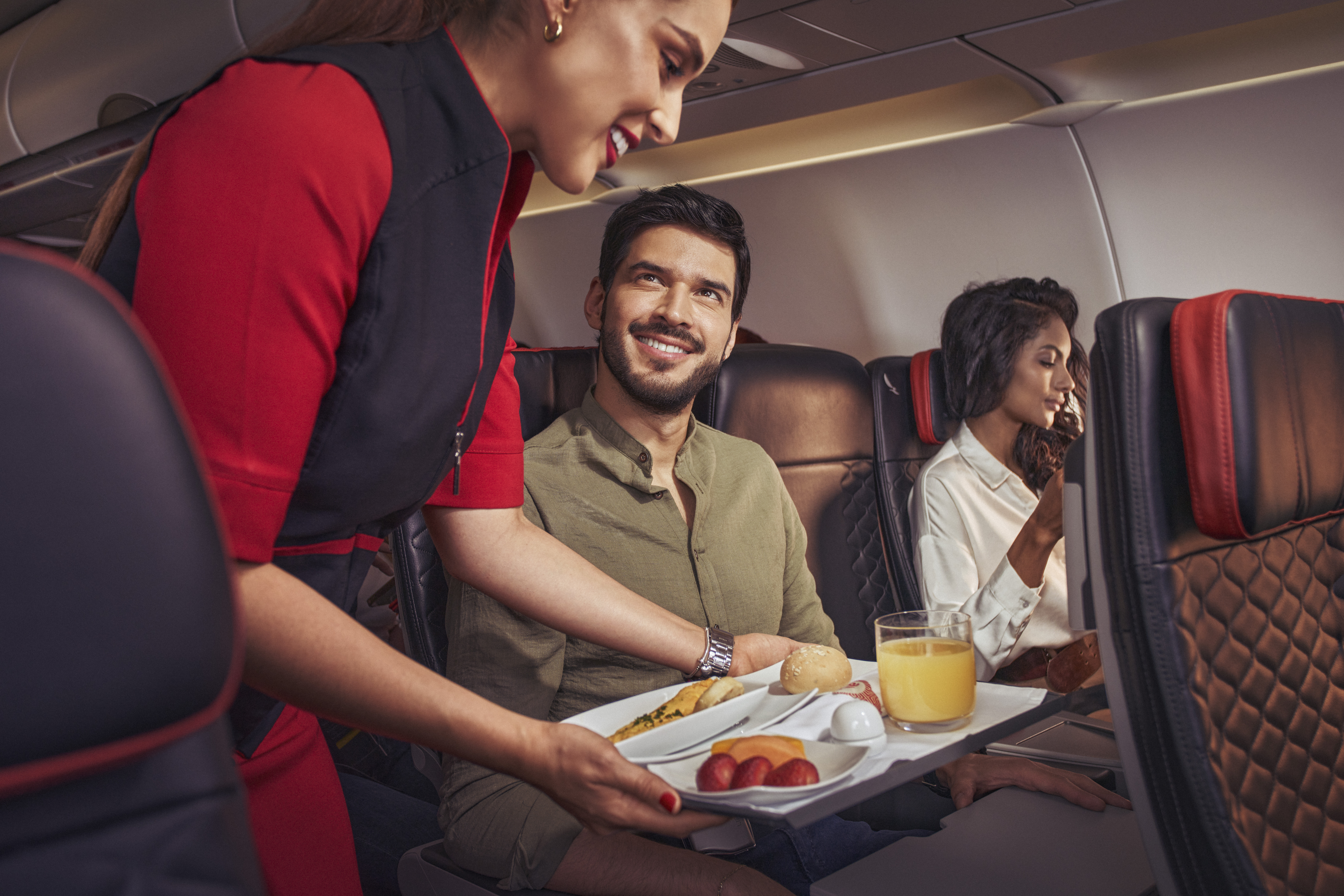 A flight attendant in a red and black uniform serves a meal tray with food and a glass of orange juice to passengers seated in an airplane.
