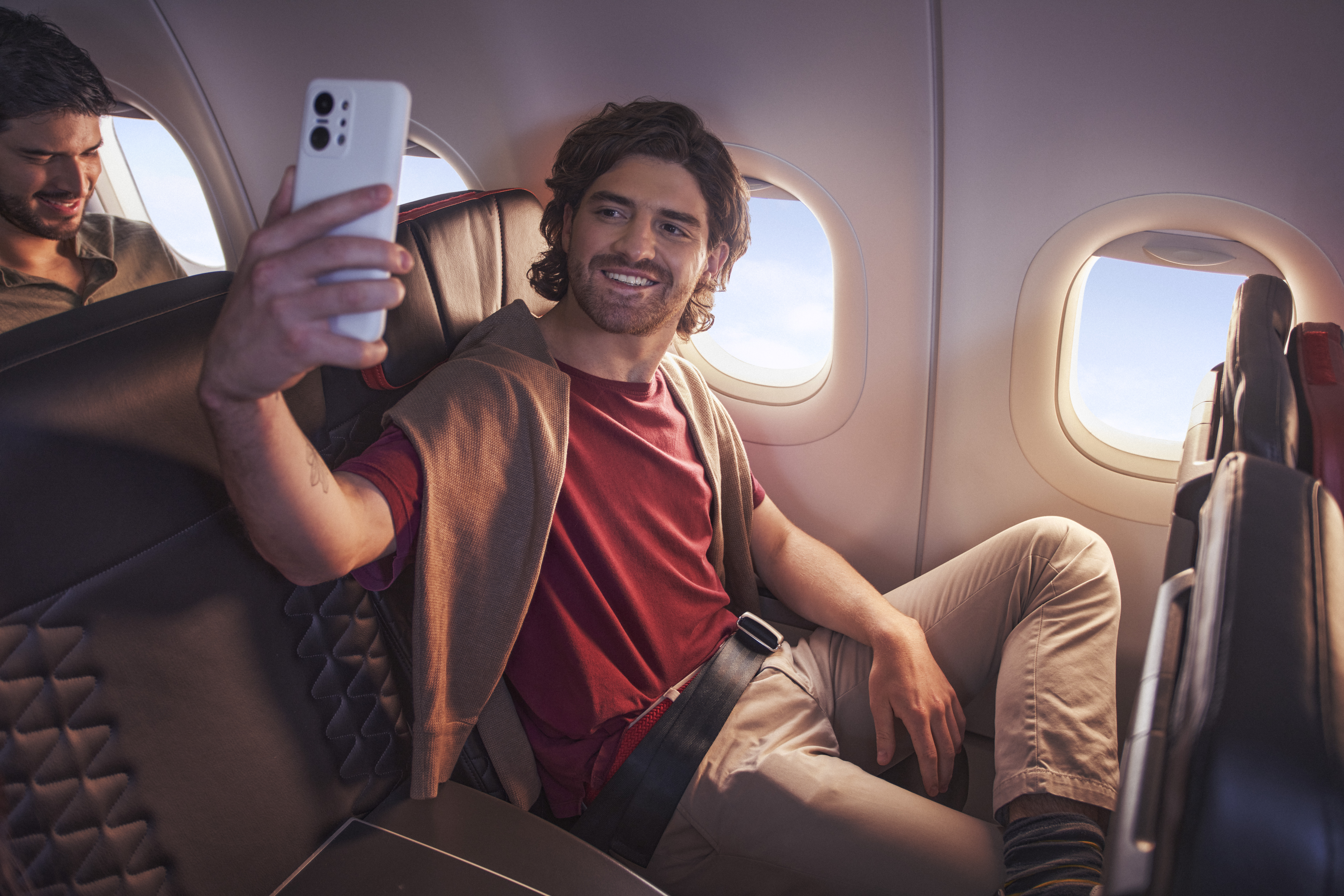  A person sitting in an airplane seat, holding a smartphone to take a selfie. The person is wearing a red shirt and beige pants, with a sweater draped over their shoulders. The airplane window shows a clear sky outside.