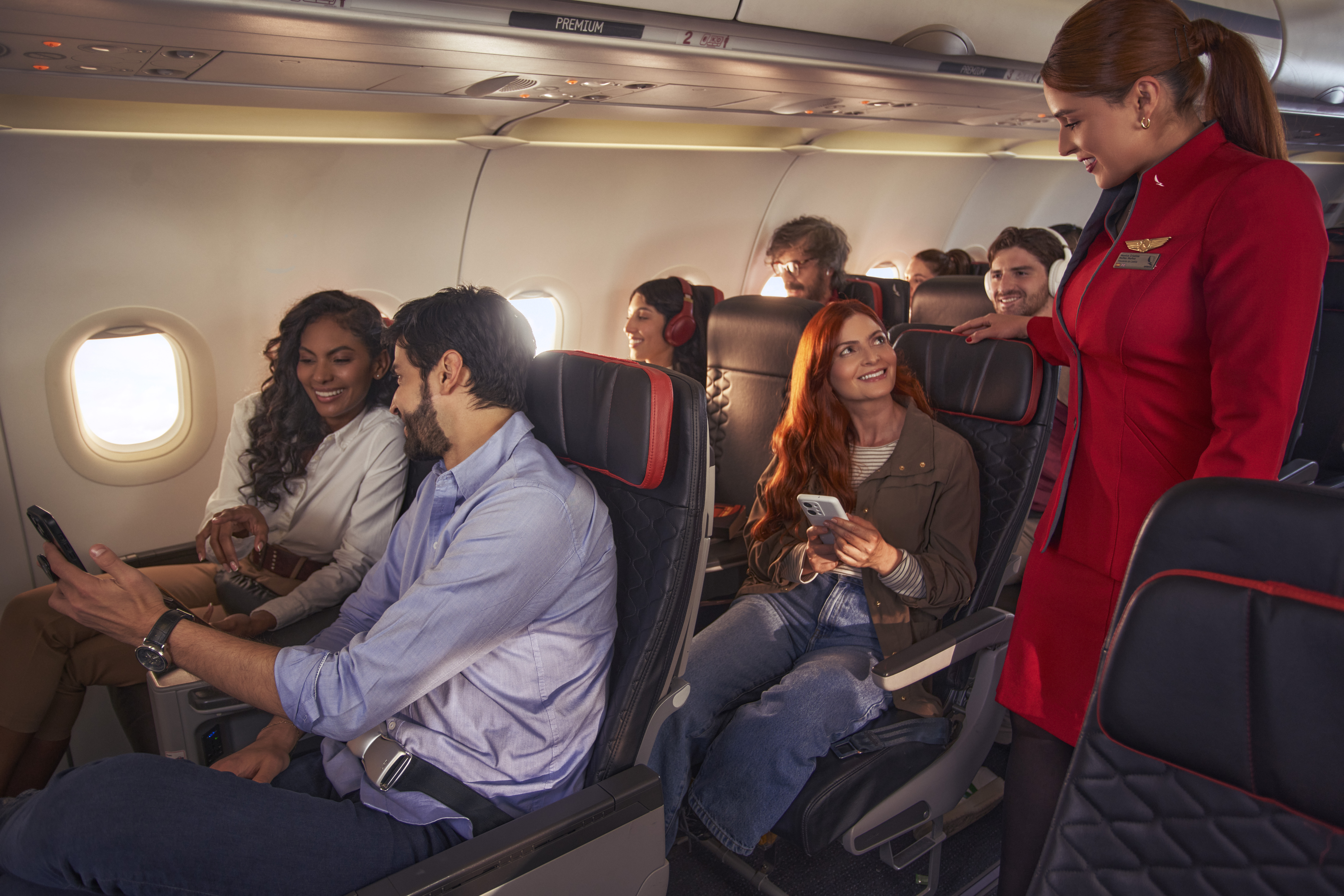 A group of passengers seated in an airplane cabin, using their mobile phones. A flight attendant in a red uniform is standing and assisting one of the passengers. The interior of the airplane shows rows of seats and windows.