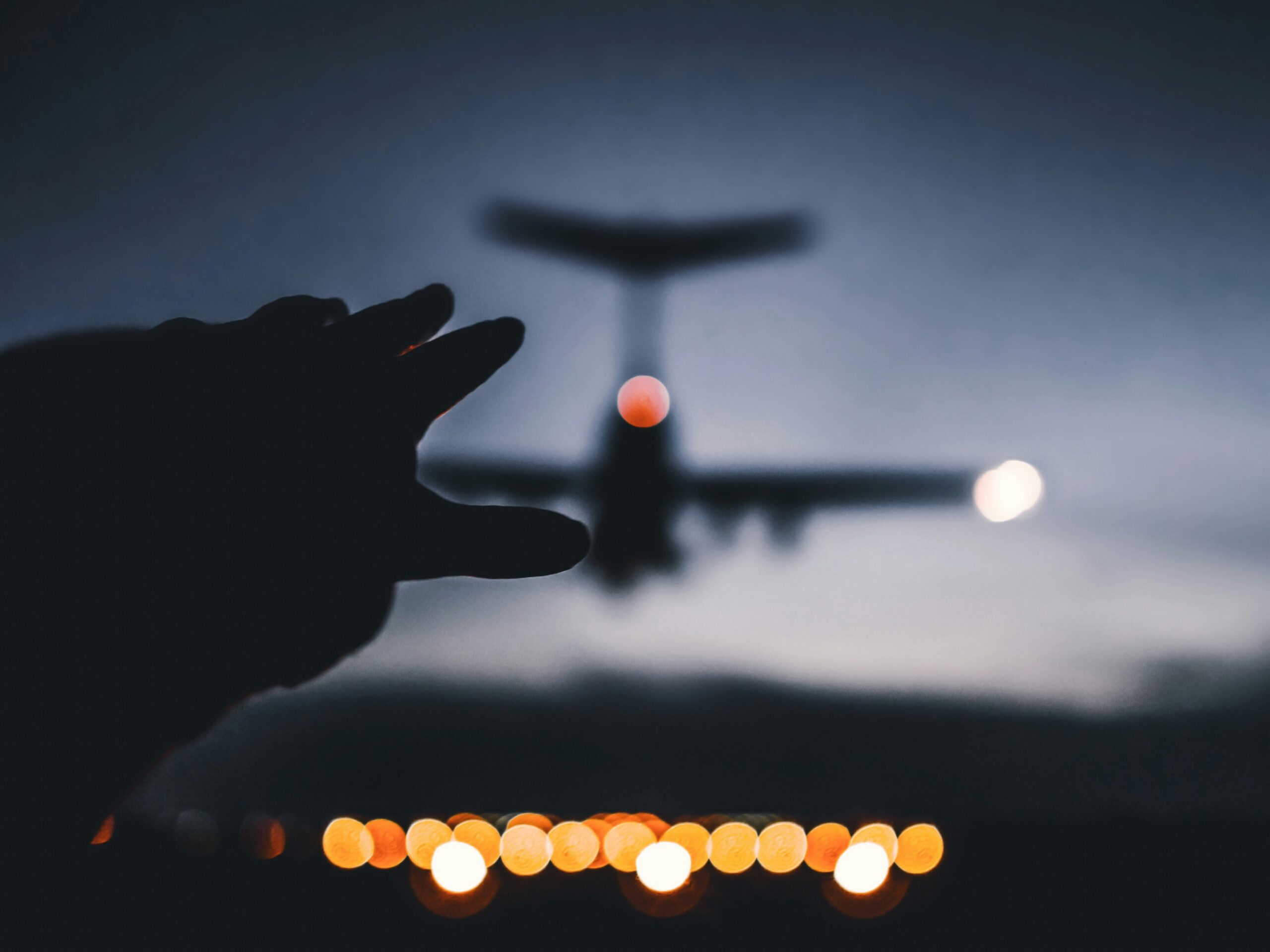 A silhouette of a hand reaching towards an airplane flying overhead at dusk, with blurred city lights in the background.