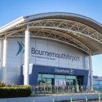 The front entrance of Bournemouth Airport, featuring a large arched roof and the main sign that reads "Bournemouth Airport - Part of Regional & City Airports." Below the sign is an entrance labeled "Departures" with glass doors and various signs indicating directions and restrictions.