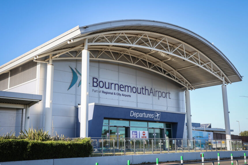 The front entrance of Bournemouth Airport, featuring a large arched roof and the main sign that reads "Bournemouth Airport - Part of Regional & City Airports." Below the sign is an entrance labeled "Departures" with glass doors and various signs indicating directions and restrictions.