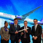 A group of five people stand in front of a large screen displaying an image of an Avelo Airlines airplane flying against a blue sky with clouds. The individuals are dressed in formal and professional attire, with two men in suits holding a model airplane.