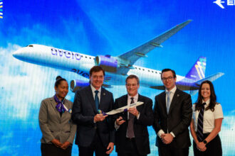 A group of five people stand in front of a large screen displaying an image of an Avelo Airlines airplane flying against a blue sky with clouds. The individuals are dressed in formal and professional attire, with two men in suits holding a model airplane.
