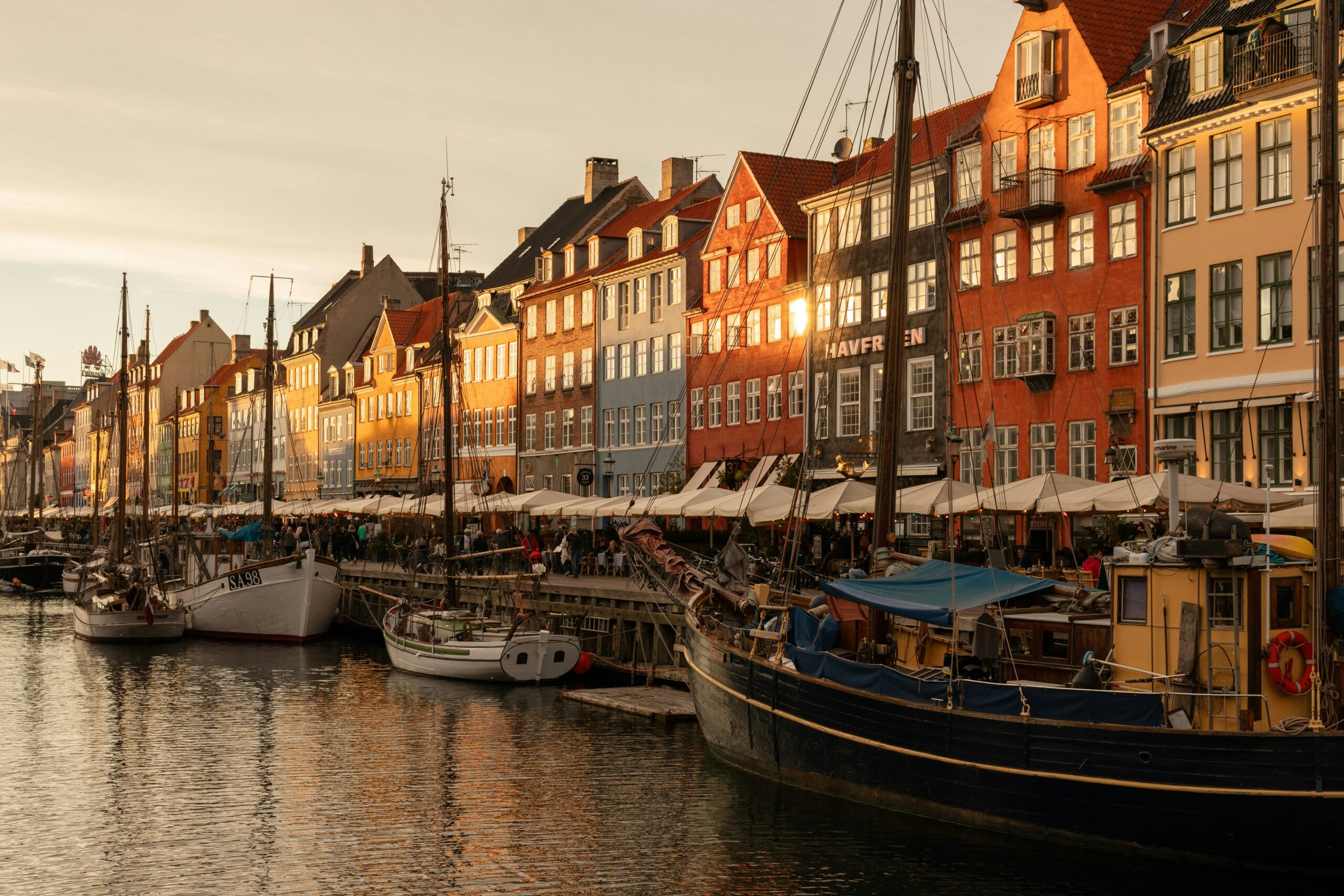 A picturesque view of Nyhavn, Copenhagen during sunset. The image captures colorful historic buildings lining the waterfront, with several boats moored along the canal. The warm sunlight casts a golden glow on the facades of the buildings and reflects off the water, creating a serene and inviting atmosphere. People can be seen walking along the promenade under white awnings.