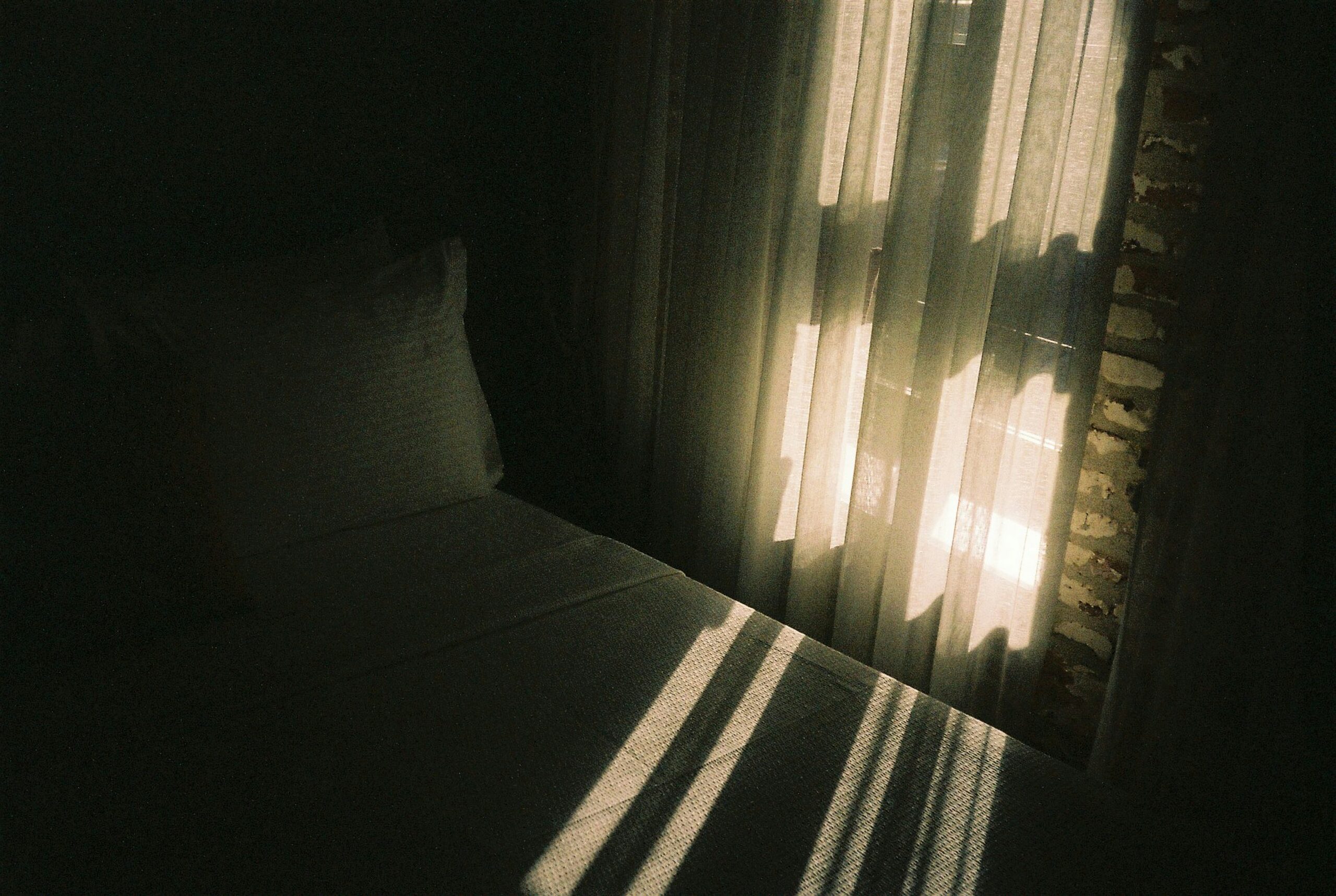 dimly lit bedroom with sunlight streaming through sheer curtains, casting shadows on a neatly made bed with white linens and a pillow. The light creates a serene and tranquil atmosphere in the room.